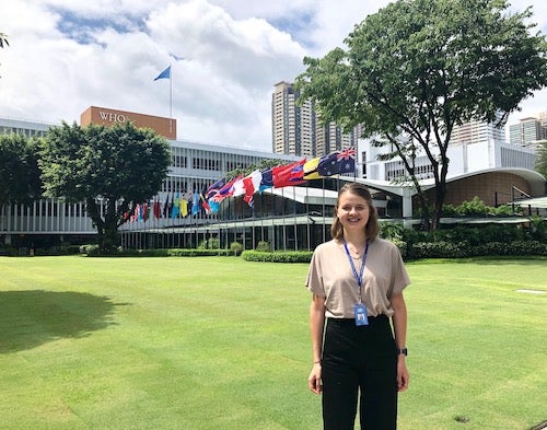 Emily Graul (NHS’20) stands on a lawn in front of the flags of various countries near a World Health Organization building.