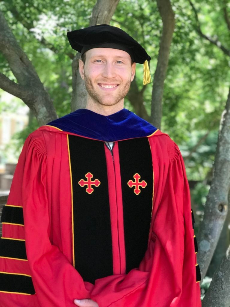 Dr. Paul Jacob “Jake” Bueno de Mesquita (NHS’12) in his doctoral regalia including red gown and cap standing in front of trees