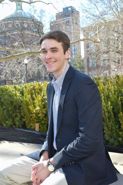 Matthew Chakwin (NHS’21), wearing business attire, sits down in front of plants and buildings.