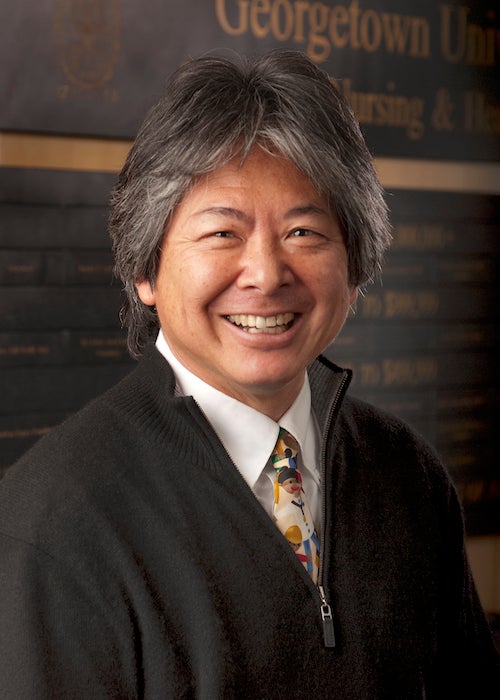 Dr. Martin Iguchi in a portrait-style photograph in St. Mary's Hall in front of the donor wall with the word Georgetown visible.