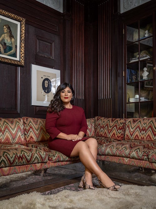 Vanessa Villaverde sits for a formal photograph on a couch with paintings and a shelf with books and statues behind her.