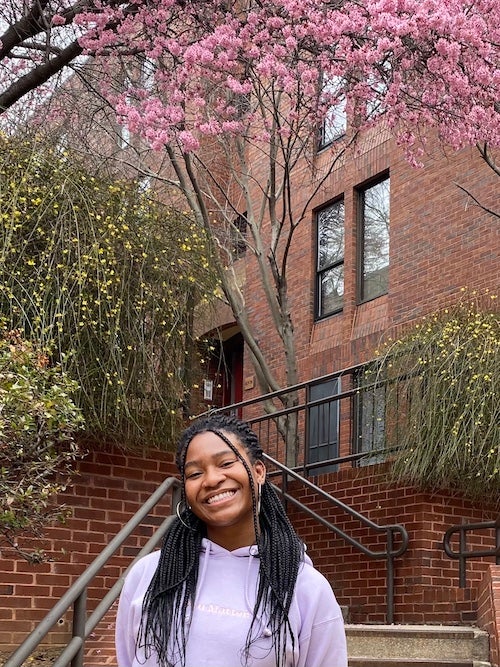Melody Emenyonu stands on Georgetown's campus in front of a residence hall and trees in blossom.
