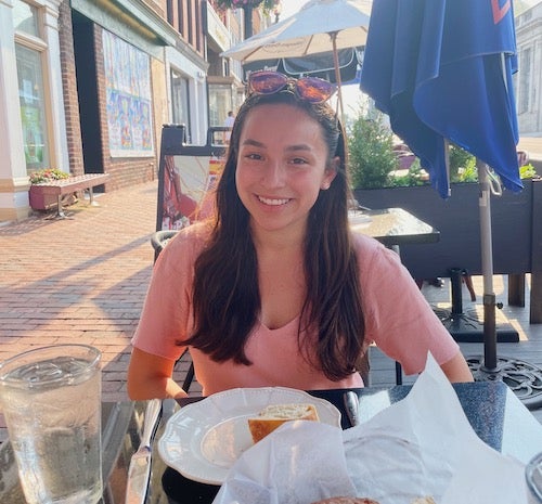 Lizzie Graham, a human science major, sits at an outdoor restaurant along a city street with an umbrella and buildings behind her and a set table in front of her.