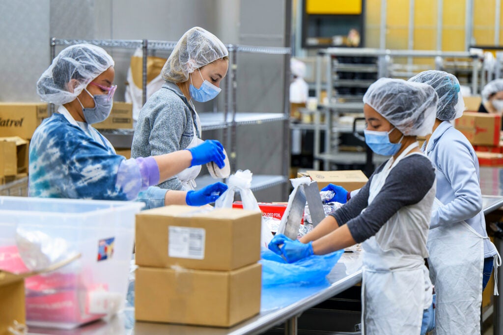 Students garbed in protective equipment pack food at a large table