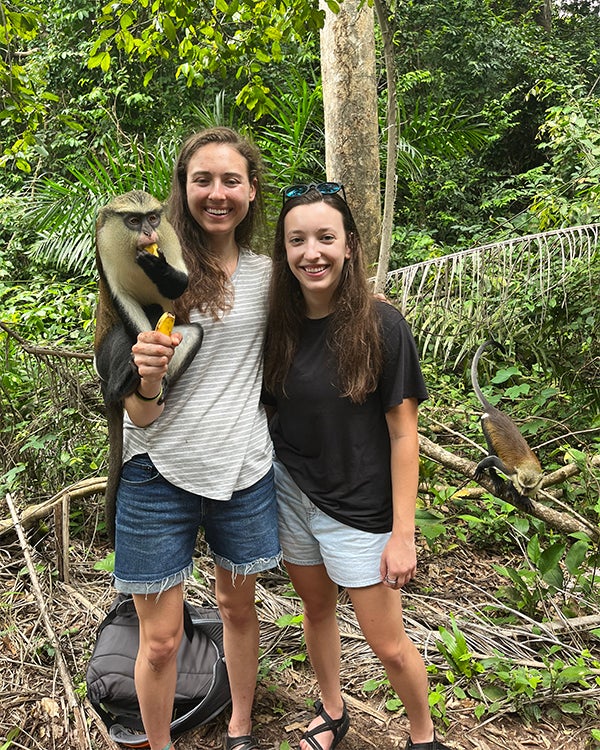 Two women stand side by side in a jungle area, one has a monkey perched on her shoulder eating a banana