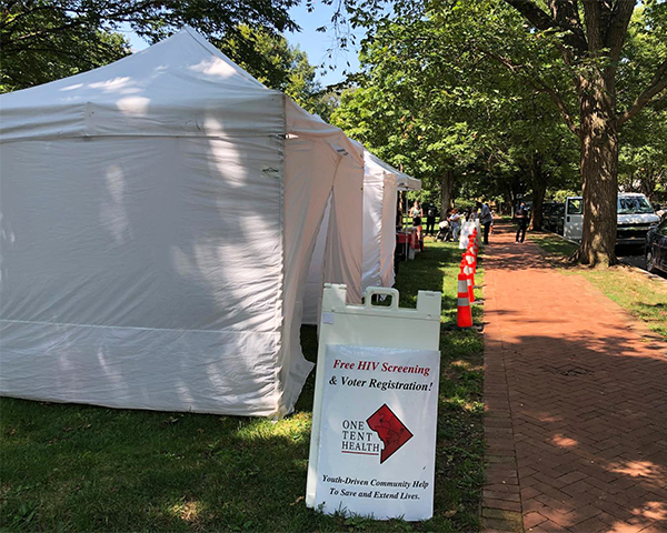Two large white canvas tents occupy space near a brick sidewalk, a One Tent Health sign is in front of them
