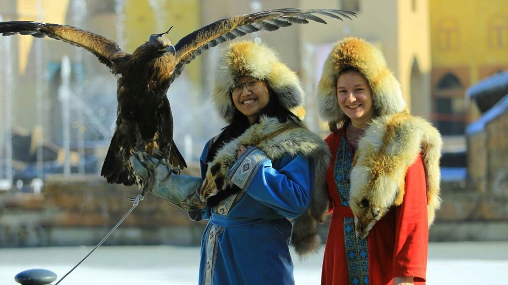 Two women stand draped in furs, one holding an eagle with wings outstretched