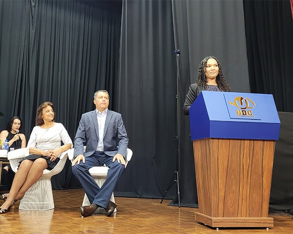 A young woman stands at a podium with others seated behind her