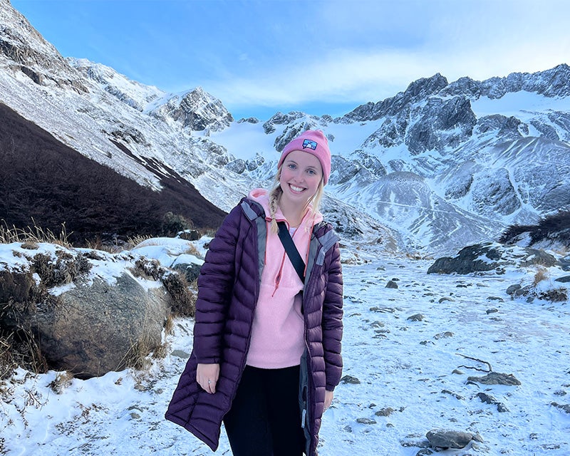 Charlotte stands in hat and parka before a snowy slope and mountain range