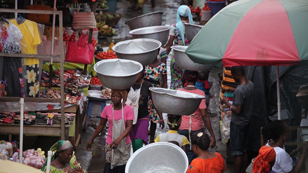 A market scene in Africa in which women carry large metal buckets on their heads
