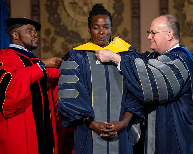 Dean King and President DeGioia place a symbolic hood over Toyin Ajayi's shoulders
