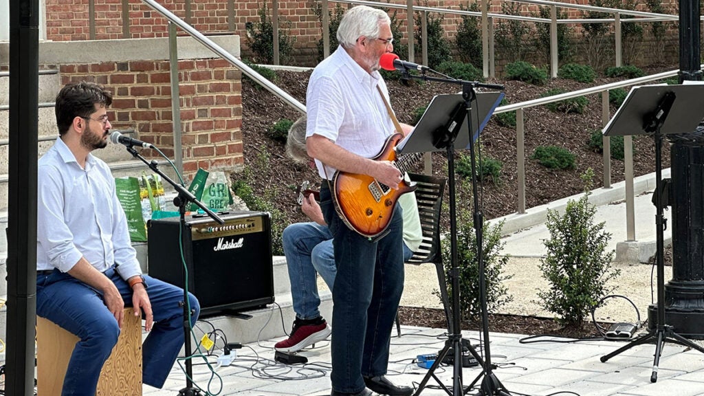 A man stands and plays a guitar while another sits on a box near a microphone; a third individual is just visible seated behind them also playing an instrument