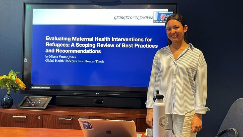 A young woman stands next to a monitor on which the words Evaluating Maternal Health Interventions for Refugees: A Scoping Review of Best Practices and Recommendations by Nicole Vernot-Jones Global Health Undergraduate Honors Thesis" are displayed