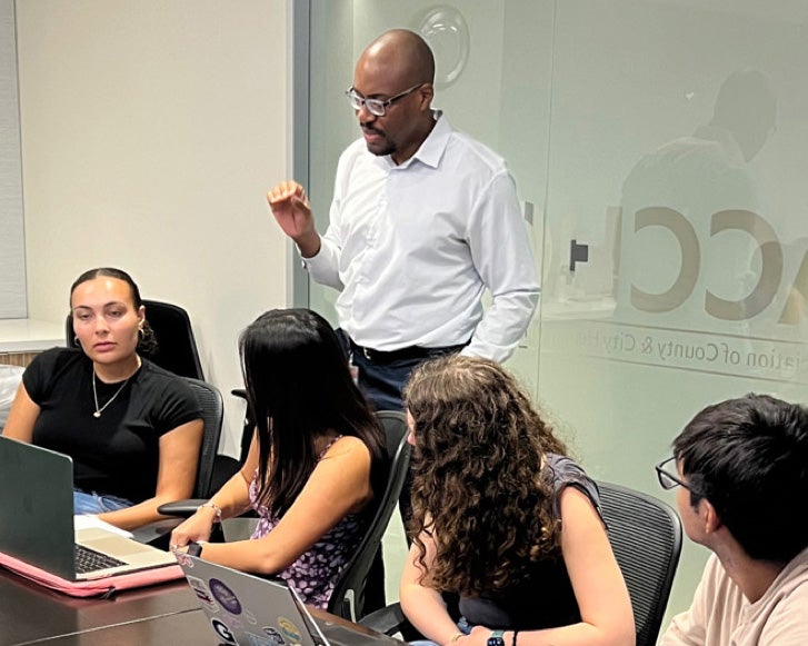 A NACCHO staff member speaks with several students who are seated at a table