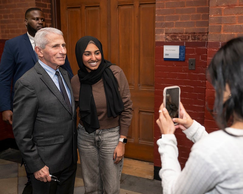 Fauci and Isra stand together while a woman takes their picture on her cellphone, a secret service agent stands in the background