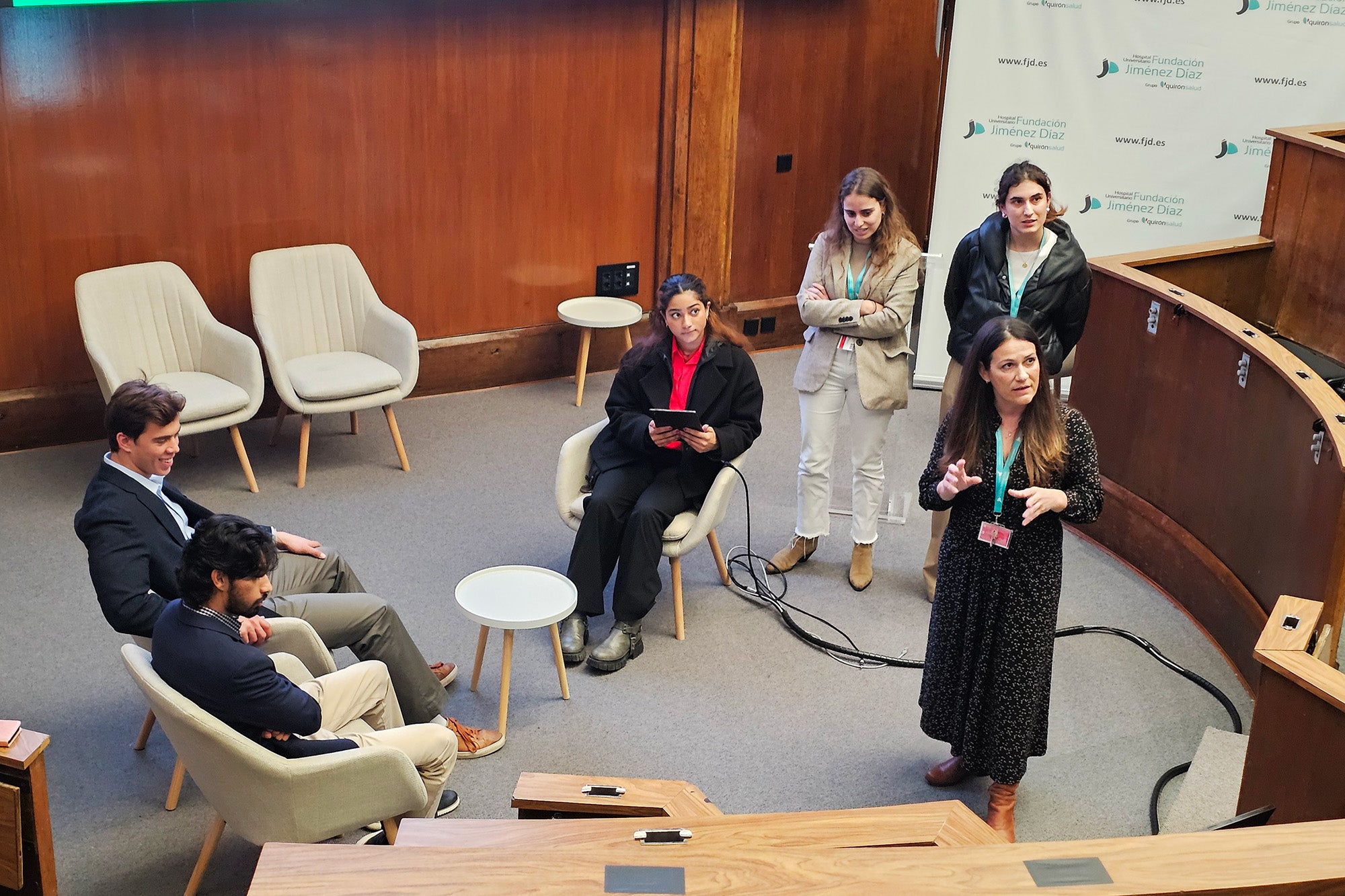 Some students sit while others stand while a woman speaks to the group at the Fundacion Jimenez Diaz