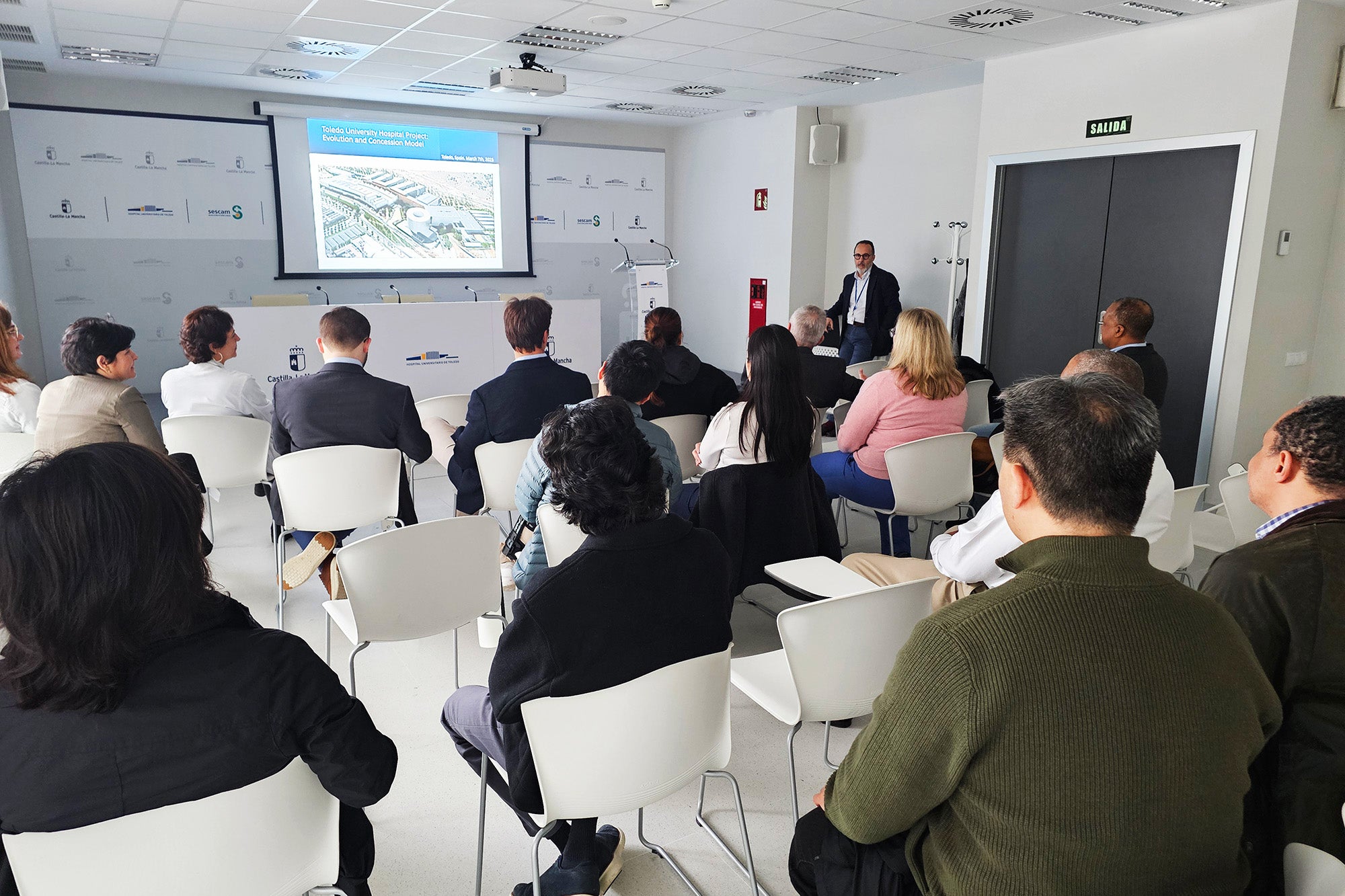 Individuals sit in chairs facing a speaker and presentation projected on a screen at the front of the room