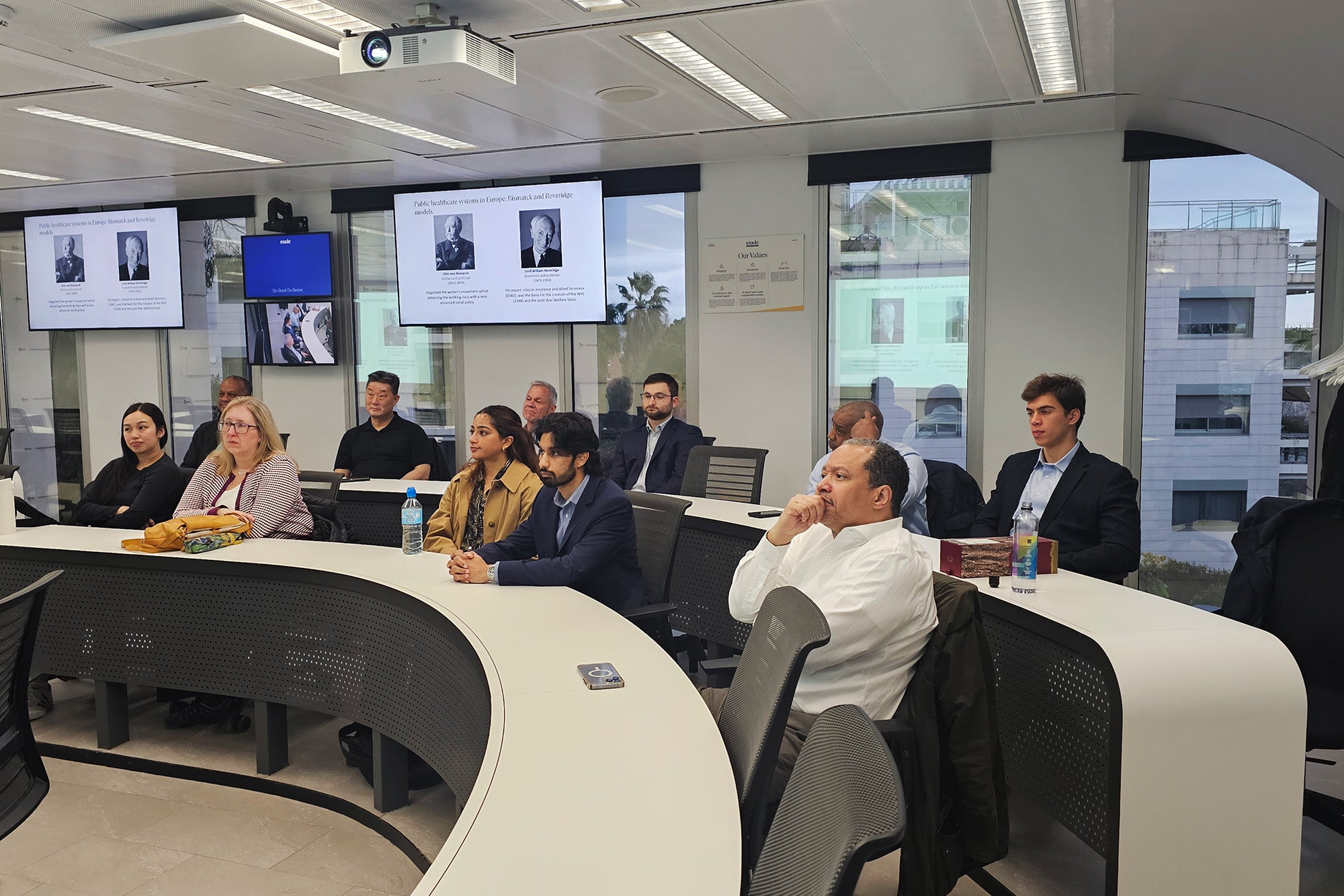 Individuals sit at long tables and listen to a speaker