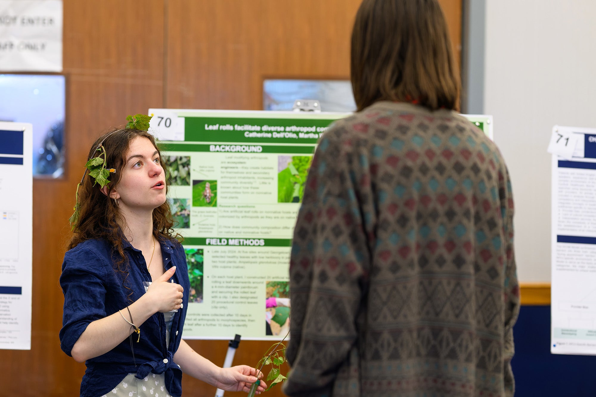 A woman wearing a garland of green ivy speaks to a person about her research poster behind her