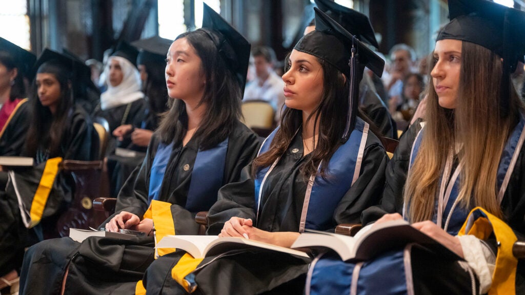 Students in caps and gowns are seated in a row and listening during Commencement