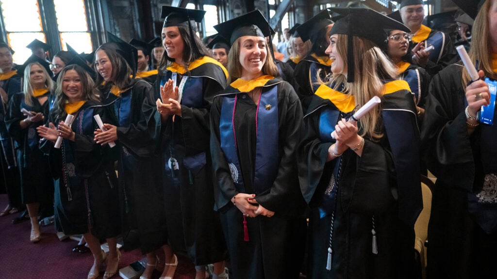 Students in caps and gowns and holding diplomas stand in a line at Commencement