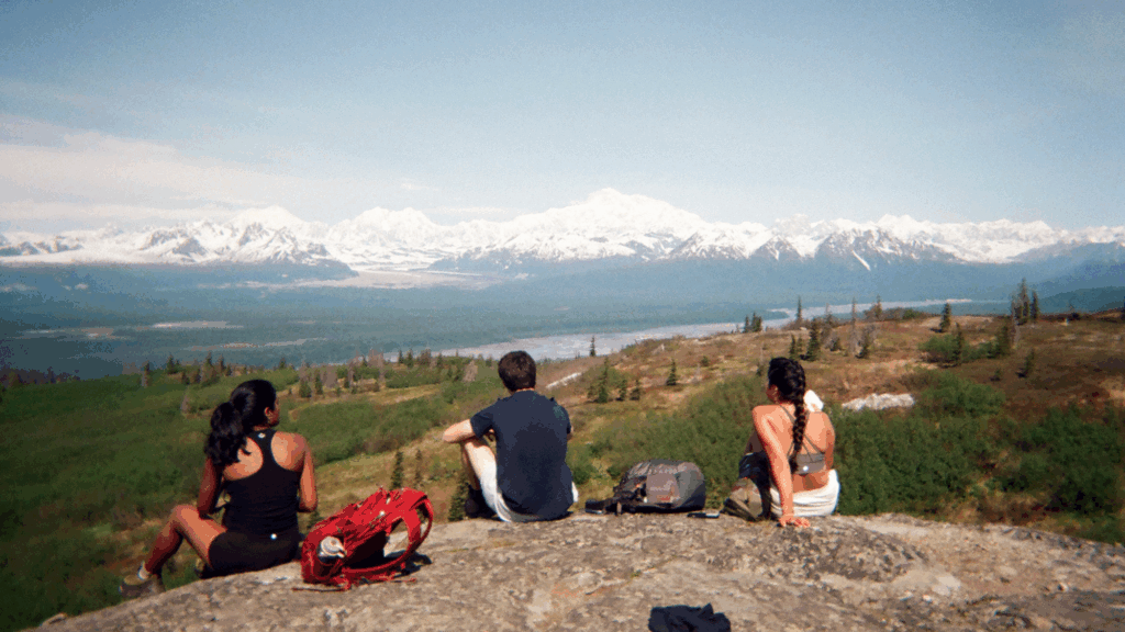 Three students sit on a rocky outcrop and contemplate a mountain vista in Alaska