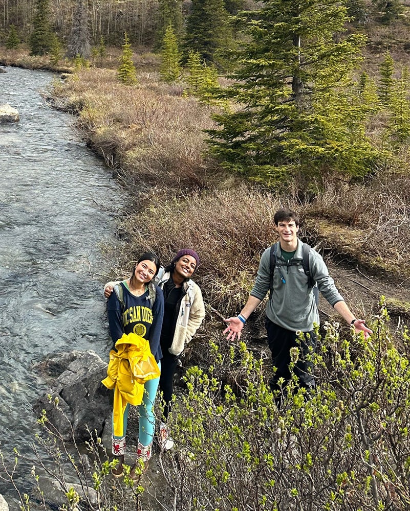 Three students stand together with a river and mountains in the Alaskan wilderness behind them