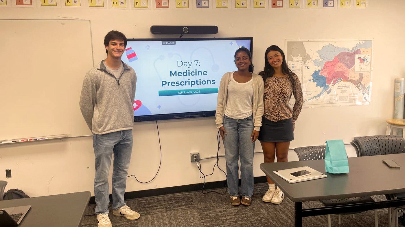 Three students stand in front of a monitor in a classroom