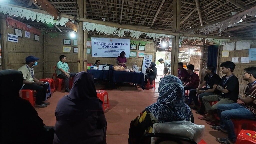 The interior of a medical clinic where people sit on chairs in a ring waiting while two people stand behind a long table at the front of the room