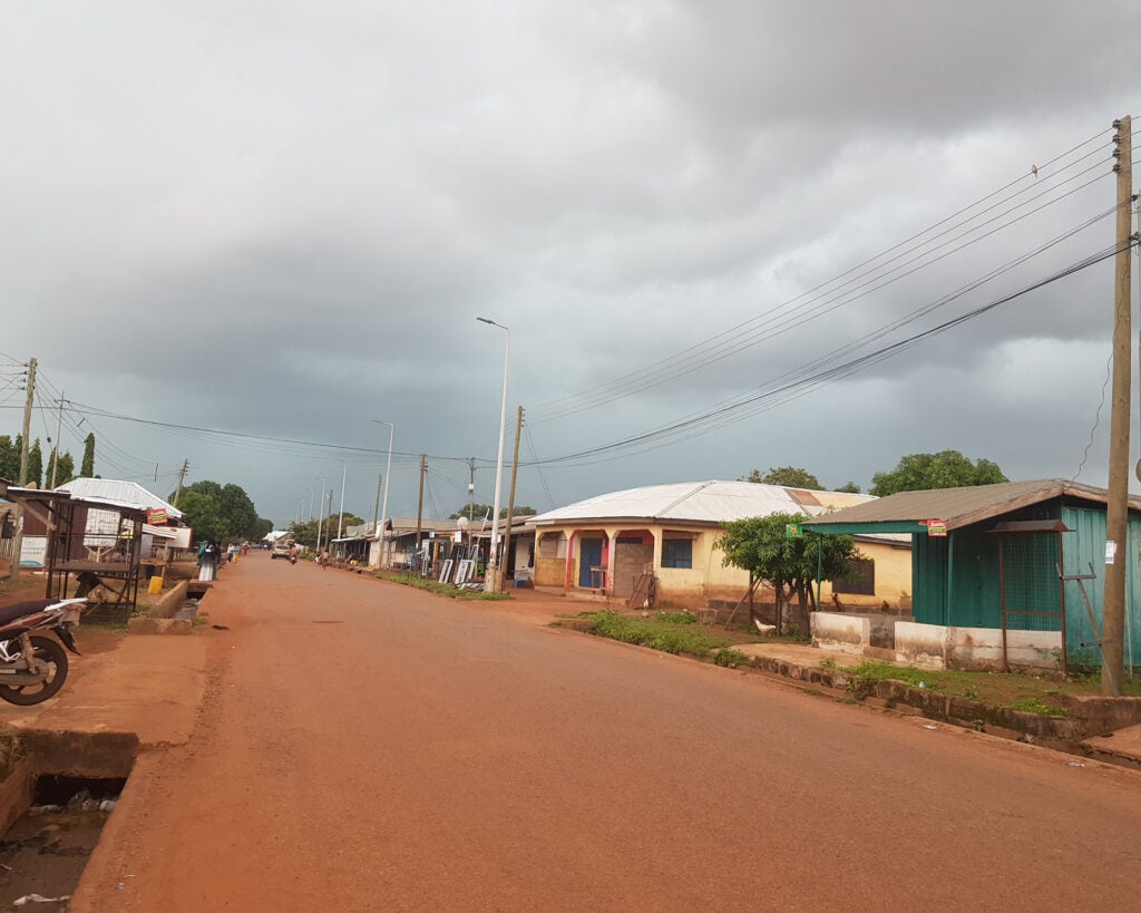 A red dirt road in Ghana lined with buildings and a sky above filled with storm clouds