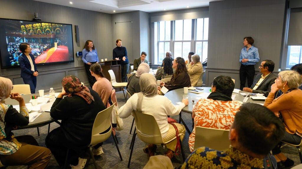 Individuals sit at tables listening to three speakers at the front of the room