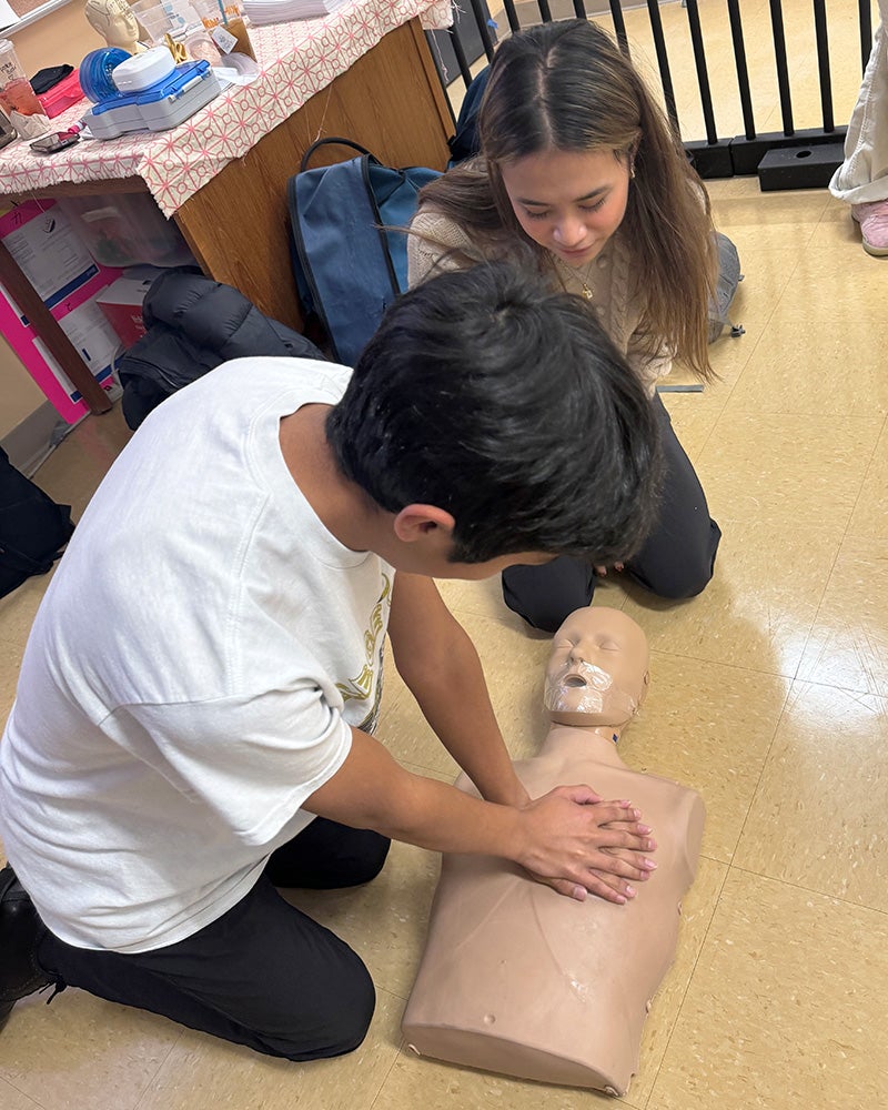 A GU student helps a high school boy learn how to administer CPR to a mannequin