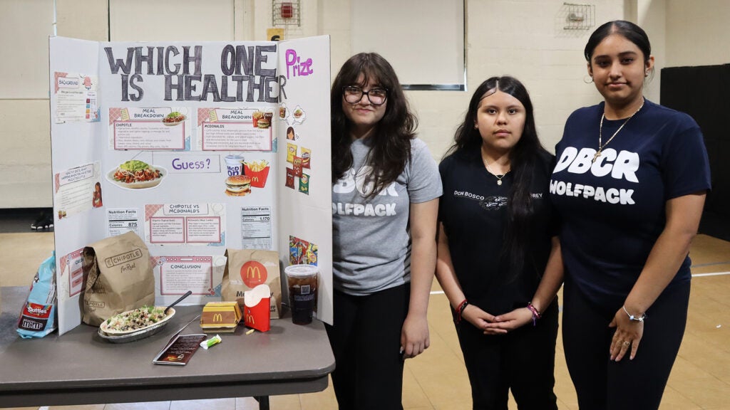 Three female high school students stand with their health fair exhibit titled Which One is Healthier? 