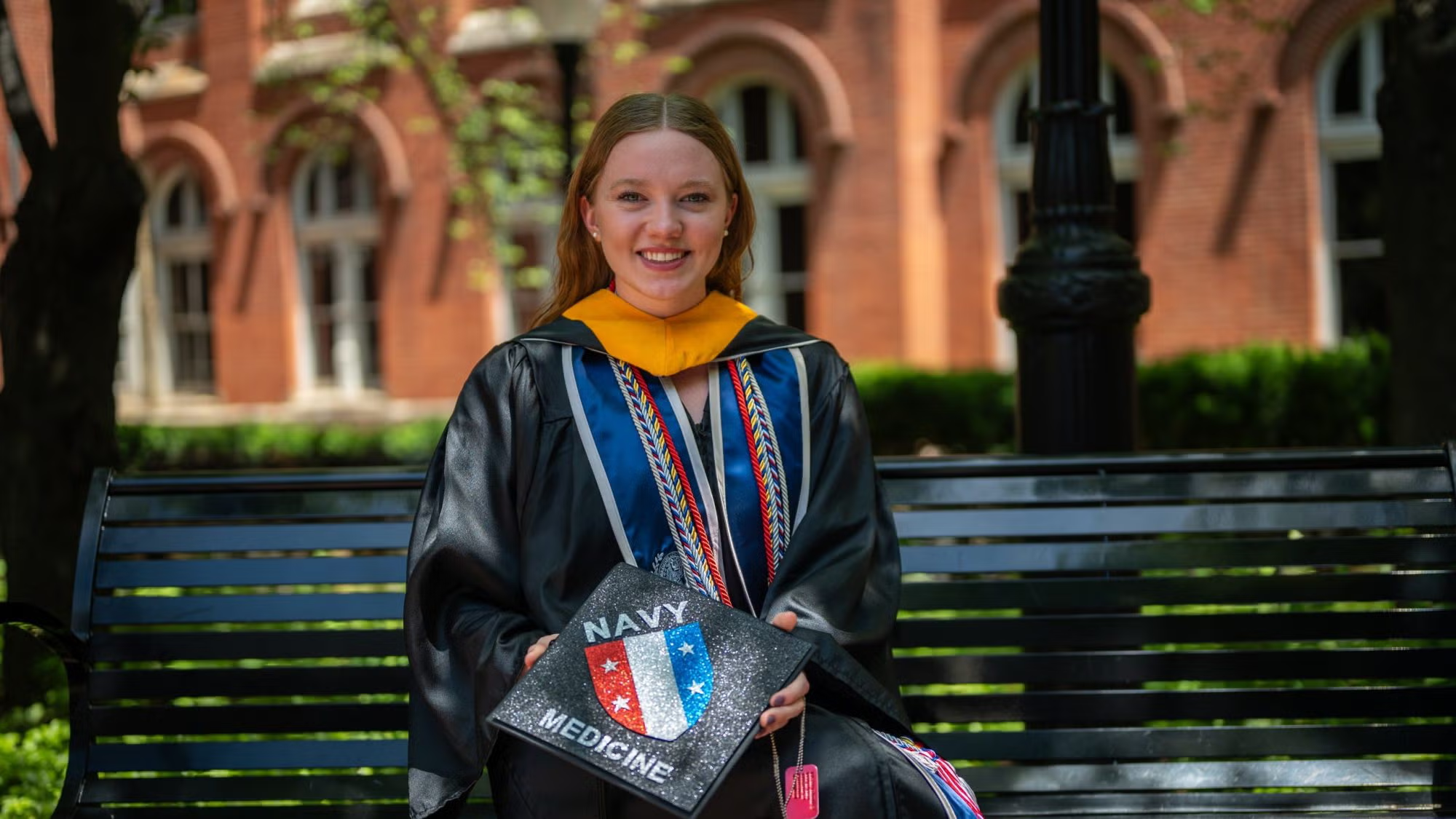 Alina Watson is dressed in cap and gown and seated on a bench on campus